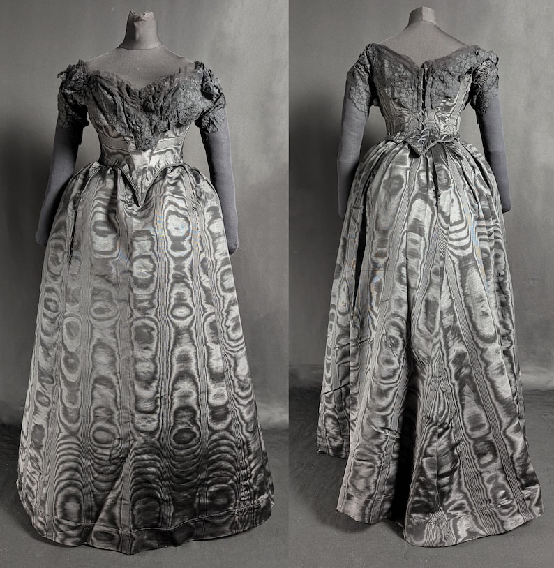 May include: A vintage, floor-length gown displayed on a mannequin. The dress features a patterned skirt and a detailed bodice with short sleeves. The fabric has a unique, swirling design in shades of gray and silver, creating a textured appearance. The dress is a historical garment.
