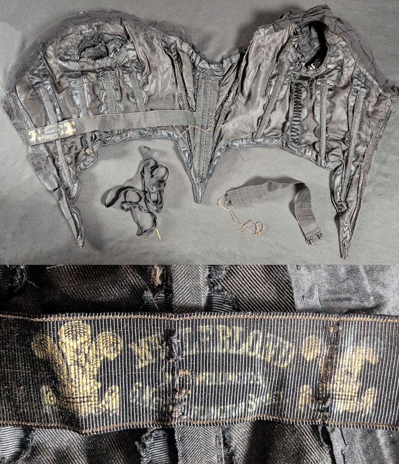 May include: A vintage black corset with boning, seams, and a label. The corset has a structured design with multiple panels and a front closure. The label features gold text and a crest. Additional black ribbons and straps are included.