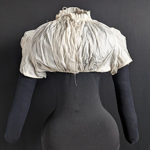 May include: A white cotton garment with a gathered neckline and short sleeves. The garment is displayed on a black mannequin.