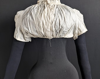 May include: A white cotton garment with a gathered neckline and short sleeves. The garment is displayed on a black mannequin.