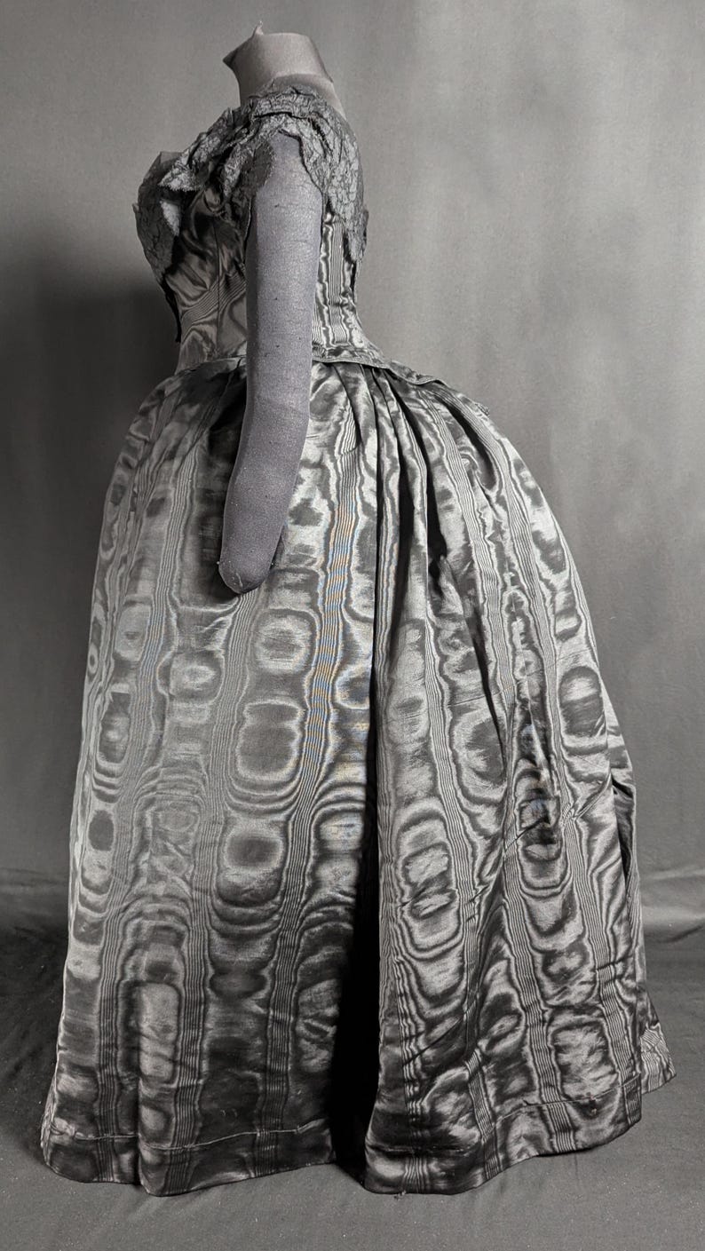 May include: A vintage gray dress with a unique pattern, featuring a fitted bodice and a full skirt. The dress has a high neckline and short sleeves. The fabric has a textured, swirling design. The dress is displayed on a mannequin.