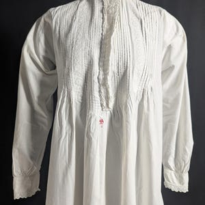 May include: A white, long-sleeved nightgown with a high, lace-trimmed collar. The bodice features vertical pleats and lace detailing down the front. The cuffs are also trimmed with lace. A small, red embroidered design is visible near the waist.