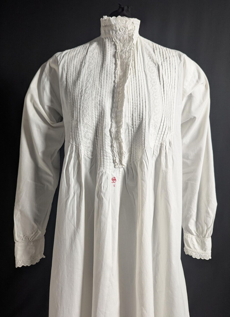 May include: A white, long-sleeved nightgown with a high, lace-trimmed collar. The bodice features vertical pleats and lace detailing down the front. The cuffs are also trimmed with lace. A small, red embroidered design is visible near the waist.