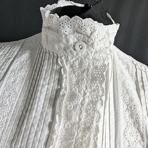 May include: Close-up of a white, vintage blouse with intricate details. The blouse features a high, ruffled collar with delicate eyelet lace trim. Vertical pleats and embroidered floral patterns adorn the front, creating a textured and elegant look. A single button secures the collar.
