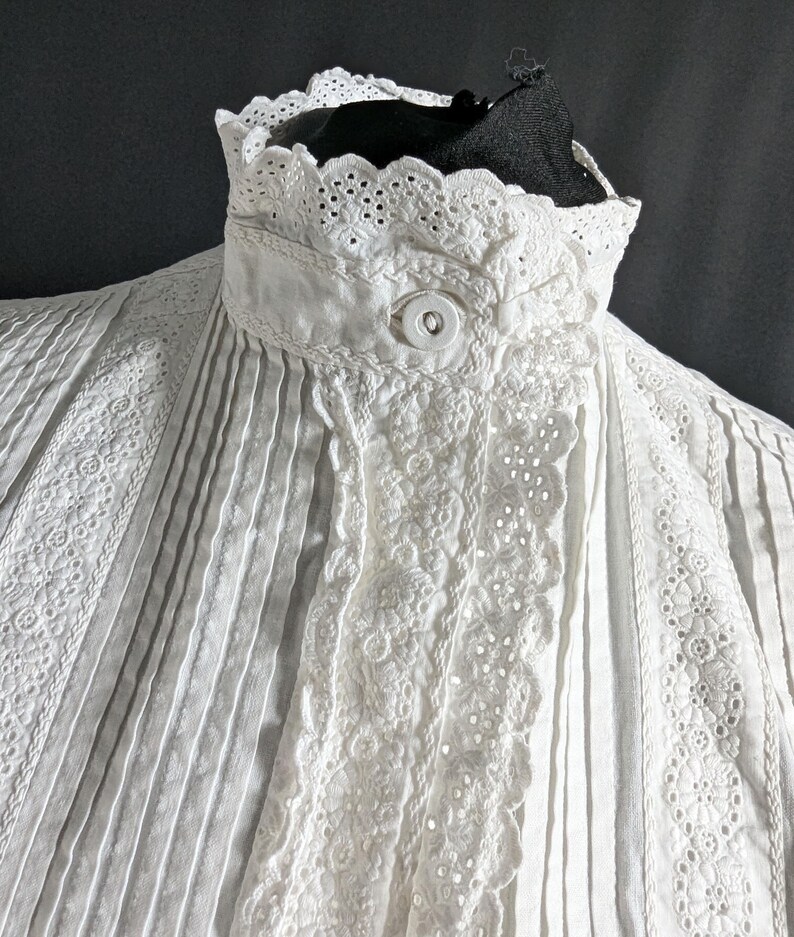 May include: Close-up of a white, vintage blouse with intricate details. The blouse features a high, ruffled collar with delicate eyelet lace trim. Vertical pleats and embroidered floral patterns adorn the front, creating a textured and elegant look. A single button secures the collar.