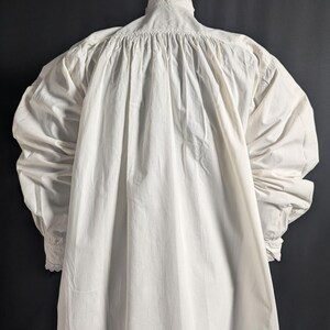 May include: A white, long-sleeved garment with a high, ruffled collar and gathered detailing at the shoulders. The sleeves are full and end in cuffs with lace trim. The fabric appears to be a lightweight cotton or linen.