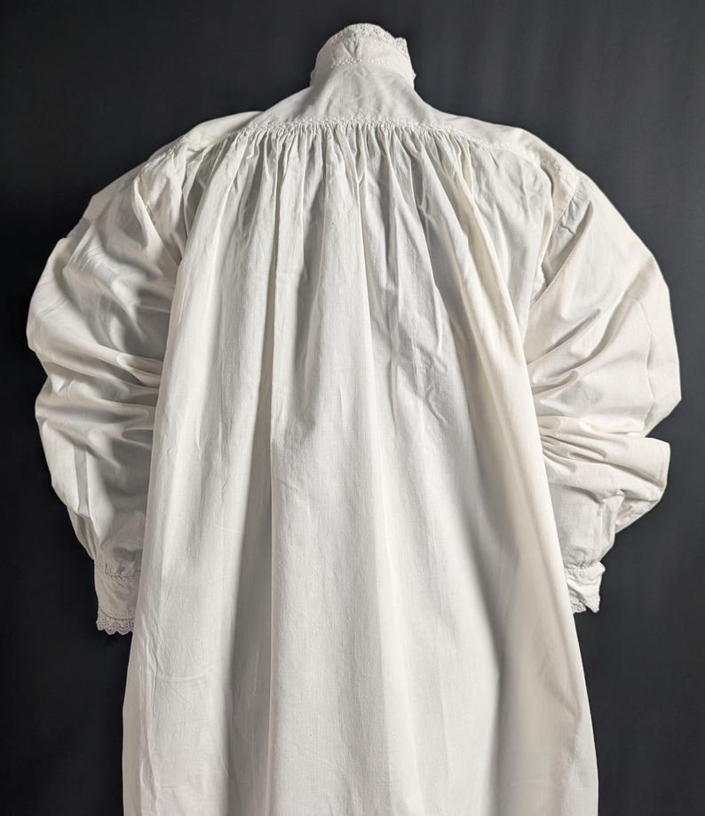 May include: A white, long-sleeved garment with a high, ruffled collar and gathered detailing at the shoulders. The sleeves are full and end in cuffs with lace trim. The fabric appears to be a lightweight cotton or linen.