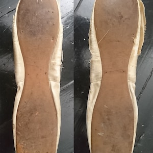 May include: A pair of white satin ballet shoes with worn leather soles. The shoes are slightly frayed around the edges.