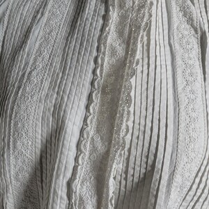 May include: Close-up of a white garment with vertical pleats and intricate lace detailing. The garment features decorative lace trim along the edges and center, creating a textured and elegant appearance. The fabric appears to be cotton or a similar lightweight material.
