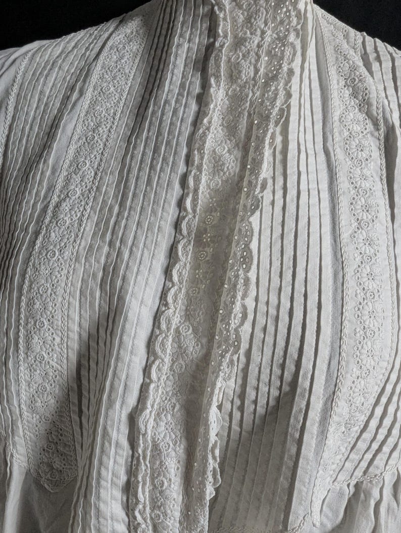 May include: Close-up of a white garment with vertical pleats and intricate lace detailing. The garment features decorative lace trim along the edges and center, creating a textured and elegant appearance. The fabric appears to be cotton or a similar lightweight material.