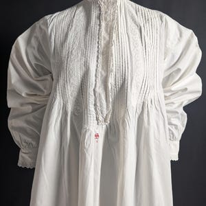 May include: A white, long-sleeved nightgown with a high, ruffled collar and button closure. The bodice features vertical pleats and lace trim. The sleeves have gathered details and lace cuffs. A small red emblem is visible near the hem.