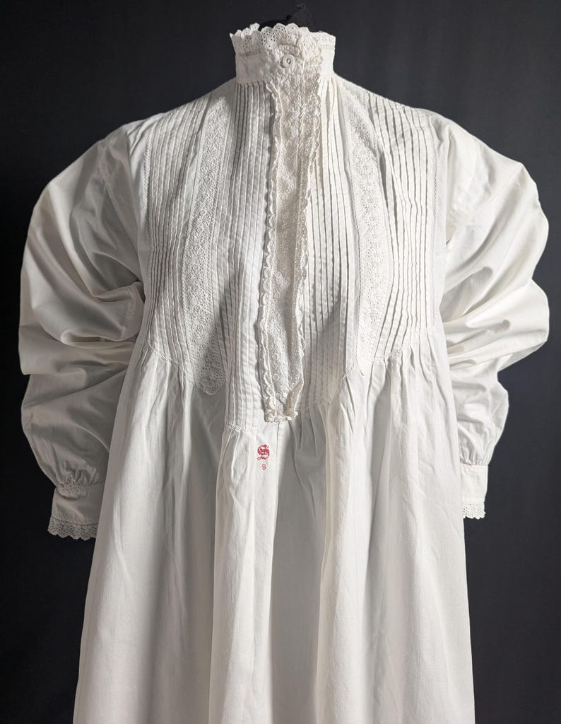 May include: A white, long-sleeved nightgown with a high, ruffled collar and button closure. The bodice features vertical pleats and lace trim. The sleeves have gathered details and lace cuffs. A small red emblem is visible near the hem.
