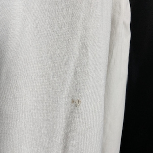 May include: Close-up of a white linen fabric with a visible weave. The fabric has a slightly textured surface and a small, light-colored imperfection. The image highlights the natural texture and color variations of the material.