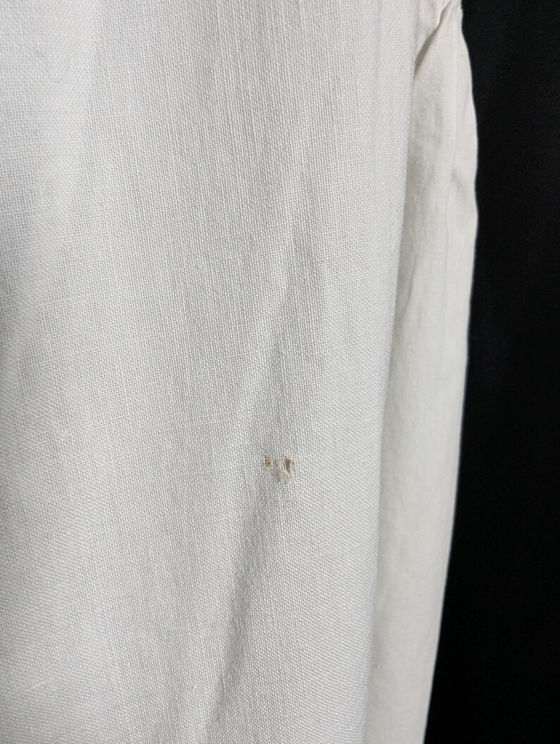 May include: Close-up of a white linen fabric with a visible weave. The fabric has a slightly textured surface and a small, light-colored imperfection. The image highlights the natural texture and color variations of the material.