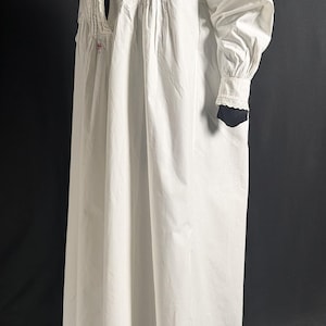 May include: A long, white nightgown with a high collar and long sleeves. The nightgown features decorative pleats and lace detailing on the bodice and cuffs. The garment is made of a lightweight fabric.