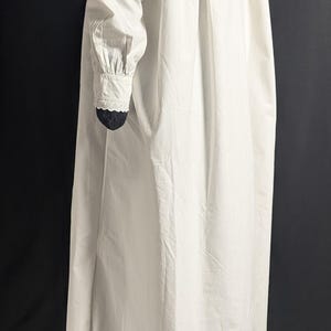 May include: A long, white nightgown with a high collar and long sleeves. The sleeves have a gathered design at the shoulder and a lace trim at the wrist. The nightgown is made of a lightweight fabric.