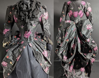 May include: A black silk dress with a floral print in shades of pink, purple, and green. The dress has a fitted bodice and a full skirt with a bustle.