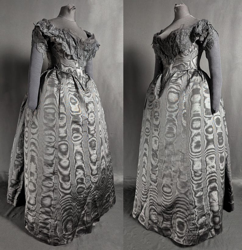 May include: A vintage gray dress with a unique pattern, displayed on a mannequin. The dress features a fitted bodice, a full skirt, and delicate lace detailing on the shoulders and sleeves. The fabric has a swirling, wood-grain-like design.