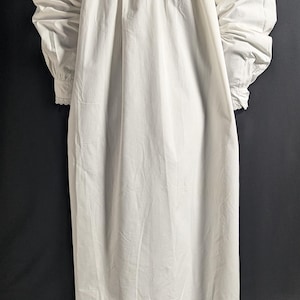 May include: A long, white nightgown with a high neckline and long sleeves. The sleeves gather at the wrists, and the gown has a loose, flowing fit. The fabric appears to be lightweight and slightly textured, with vertical folds.