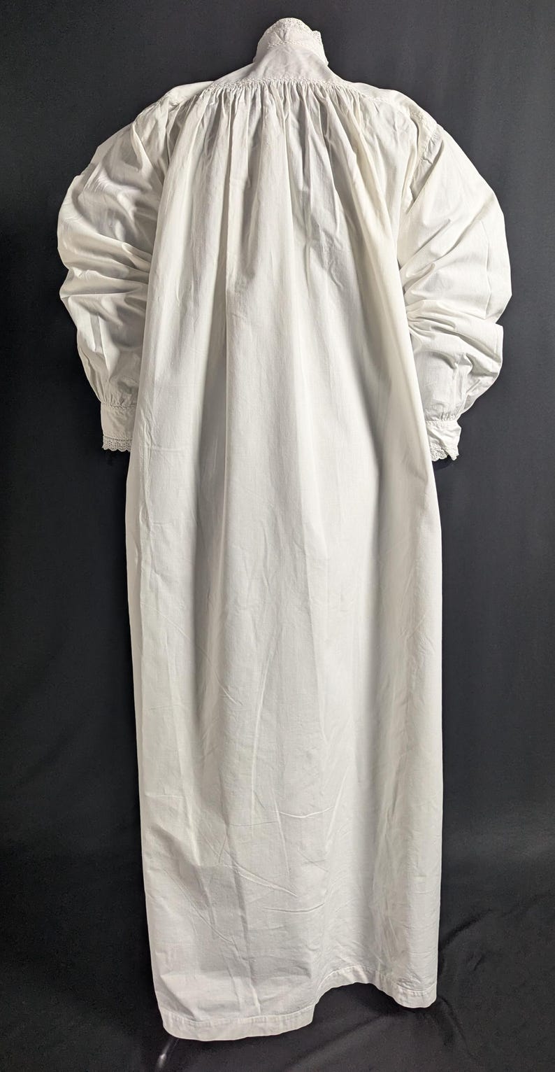 May include: A long, white nightgown with a high neckline and long sleeves. The sleeves gather at the wrists, and the gown has a loose, flowing fit. The fabric appears to be lightweight and slightly textured, with vertical folds.