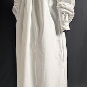 May include: A long, white cotton nightgown with long sleeves and a high, ruffled collar. The sleeves have buttoned cuffs with lace trim. The gown is gathered at the shoulders and falls to the floor.
