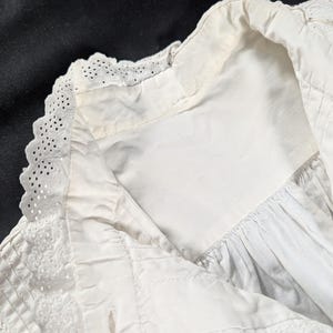 May include: Close-up of a white, vintage garment featuring intricate eyelet lace detailing along the collar and edges. The fabric appears to be cotton or linen, with a slightly textured surface. The garment has a classic, timeless design.