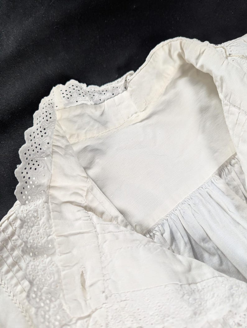 May include: Close-up of a white, vintage garment featuring intricate eyelet lace detailing along the collar and edges. The fabric appears to be cotton or linen, with a slightly textured surface. The garment has a classic, timeless design.