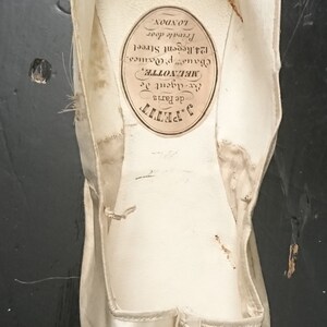 May include: A white satin ballet shoe with a label that reads "J. Petit de Paris Ex-Agent de MELNOTTE, Chaus p. Doanes. 124. Regent Street Private door LONDON."