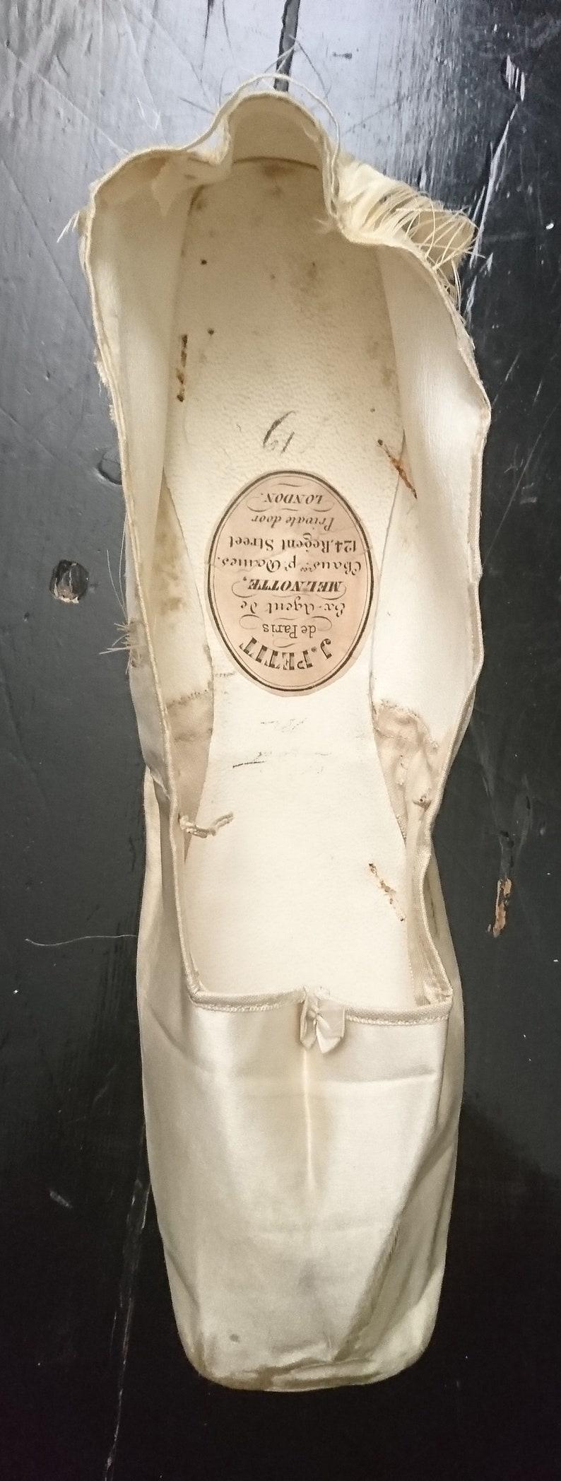 May include: A white satin ballet shoe with a label that reads "J. Petit de Paris Ex-Agent de MELNOTTE, Chaus p. Doanes. 124. Regent Street Private door LONDON."