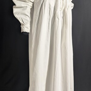 May include: A long, white nightgown with a high, ruffled collar and long sleeves. The bodice features vertical pleats and lace detailing. The sleeves have a gathered design at the shoulders and cuffs. The nightgown is made of a soft, flowing fabric.