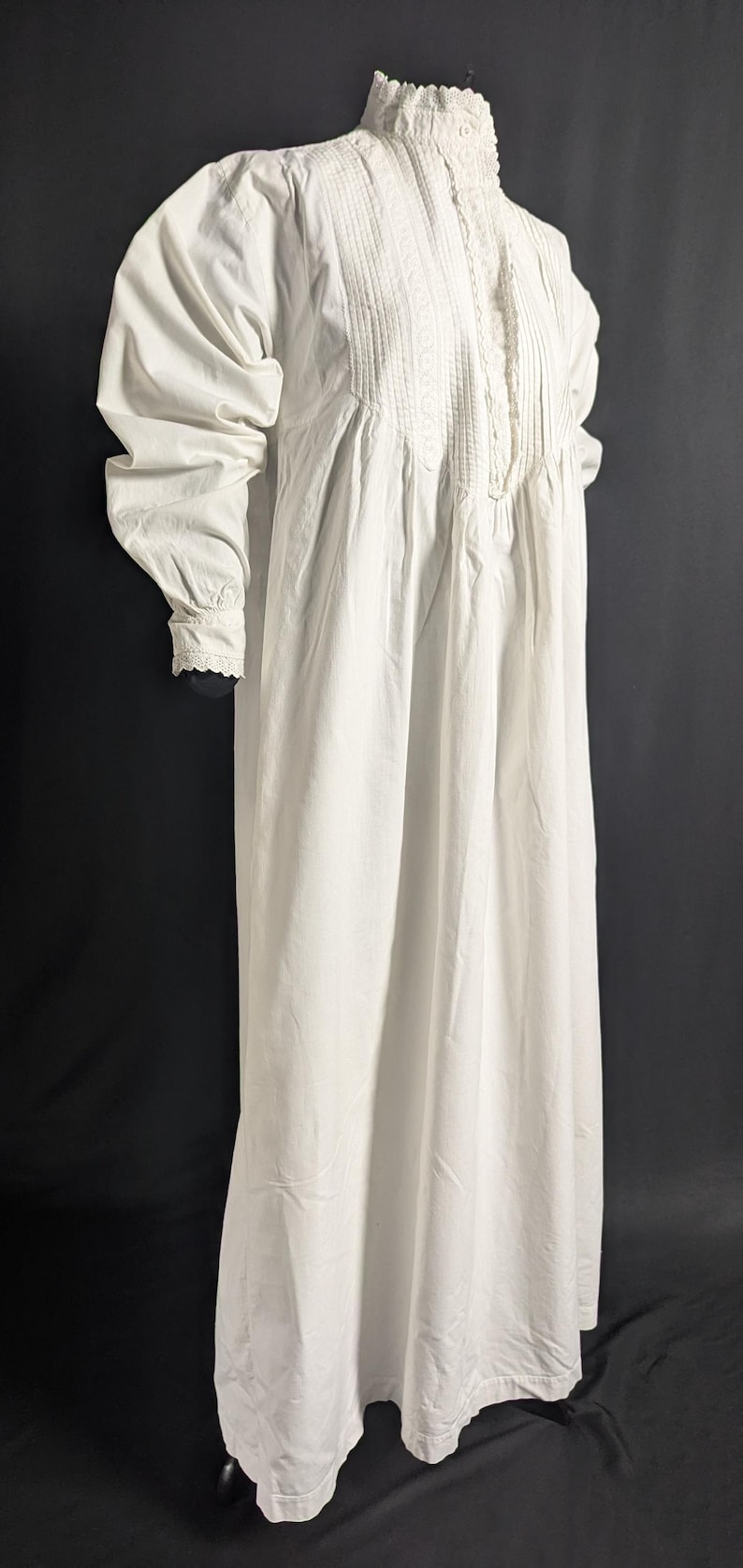 May include: A long, white nightgown with a high, ruffled collar and long sleeves. The bodice features vertical pleats and lace detailing. The sleeves have a gathered design at the shoulders and cuffs. The nightgown is made of a soft, flowing fabric.