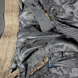 May include: Close-up of the interior of a garment, featuring black fabric with vertical stripes and a brown herringbone-patterned waistband. The garment's lining is visible, along with metal hooks and eye closures.