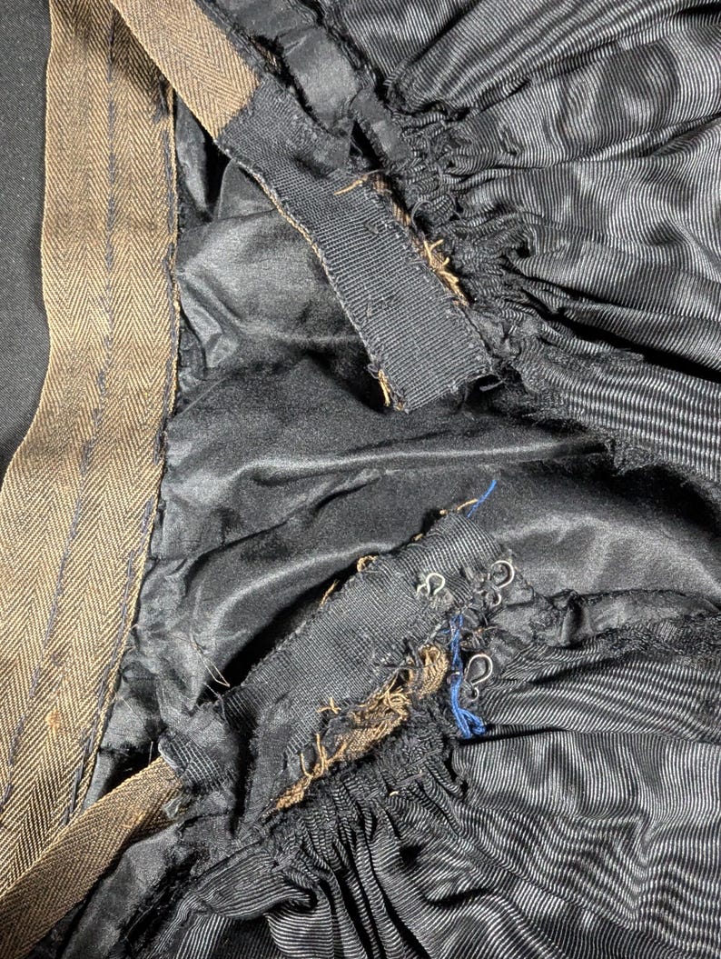 May include: Close-up of the interior of a garment, featuring black fabric with vertical stripes and a brown herringbone-patterned waistband. The garment's lining is visible, along with metal hooks and eye closures.