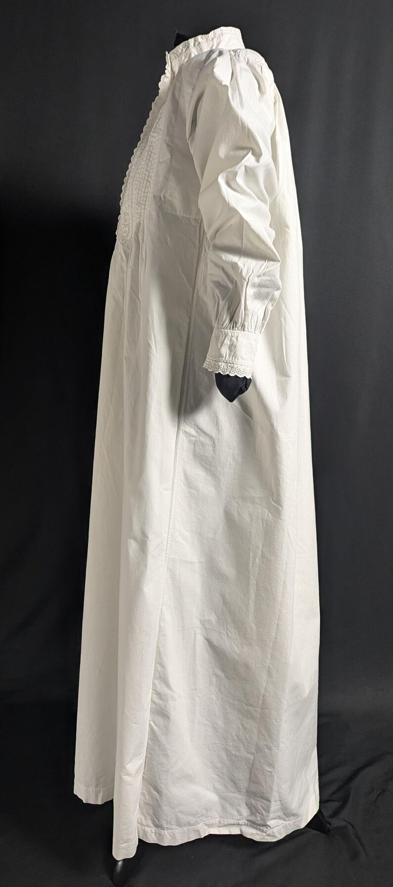 May include: A long, white nightgown with long sleeves and a high collar. The nightgown features delicate lace detailing on the collar and cuffs. The fabric appears to be a lightweight cotton or linen, suitable for sleepwear. The garment is displayed against a dark background.