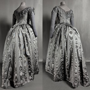 May include: A vintage gray dress with a full skirt and long sleeves, displayed on a mannequin. The dress features a patterned fabric with vertical stripes and a detailed bodice. The sleeves and bodice have a lace-like texture.