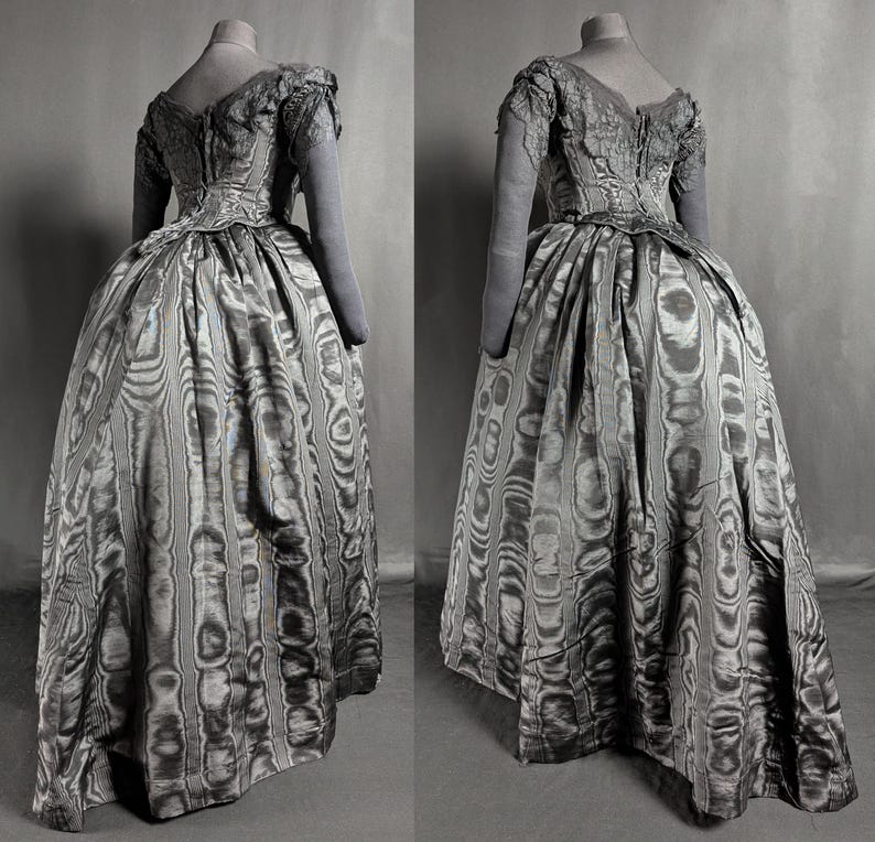May include: A vintage gray dress with a full skirt and long sleeves, displayed on a mannequin. The dress features a patterned fabric with vertical stripes and a detailed bodice. The sleeves and bodice have a lace-like texture.