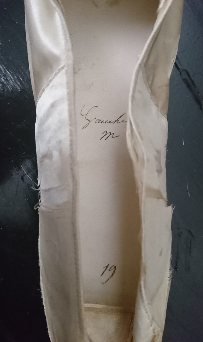 May include: A white satin ballet shoe with the inscription "Gauche" and the number "19" written inside.