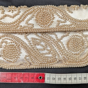 Könnte beinhalten: Elfenbeinfarbene Satin-Clutch mit aufwendiger Perlenstickerei. Die Tasche im Klappstil hat florale und wirbelnde Muster mit einem Rand aus kleinen Perlen. Ein rot-weißes Maßband liegt neben der Clutch.