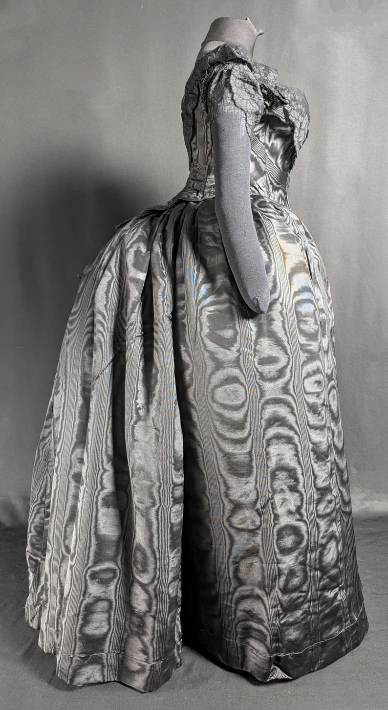 May include: A vintage-style, floor-length gown in shades of gray, featuring a unique, swirling pattern. The dress has a fitted bodice with lace detailing and short sleeves. The skirt is full and flowing, creating a dramatic silhouette. The fabric has a subtle sheen.