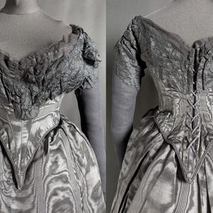 May include: A vintage gray dress with a fitted bodice and a full skirt. The dress features intricate lace detailing on the sleeves and neckline. The back of the dress has a corset-style closure. The fabric has a subtle striped pattern.
