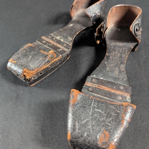Könnte beinhalten: Ein Paar antike schwarze Lederschuhe mit offenen Absätzen und Zehen. Die Schuhe haben ein geschwungenes Design mit Metallnieten und ein abgenutztes, verwittertes Aussehen. Die Innenseite ist hellbraun und die Schuhe scheinen handgefertigt zu sein.