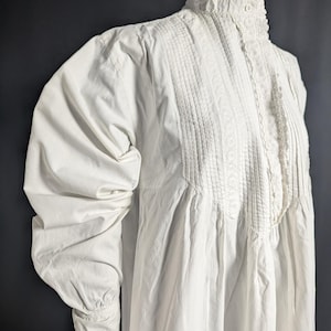 May include: A white vintage dress with a high, ruffled collar and long, puffed sleeves. The bodice features vertical pleats and delicate lace trim. The dress has a button closure at the neck and lace detailing on the cuffs.