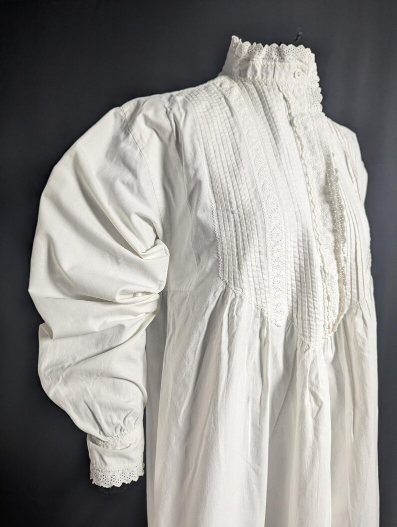 May include: A white vintage dress with a high, ruffled collar and long, puffed sleeves. The bodice features vertical pleats and delicate lace trim. The dress has a button closure at the neck and lace detailing on the cuffs.
