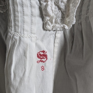 May include: Close-up of a white garment with intricate details. The fabric features vertical pleats and a delicate lace collar. A red embroidered initial "S" and the number "8" are visible on the fabric.