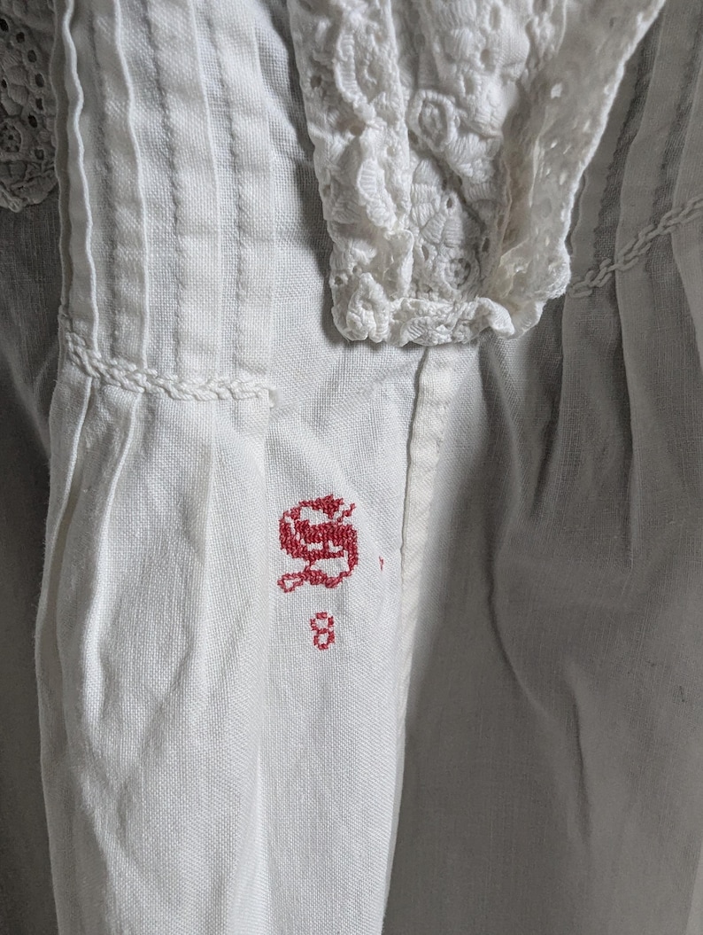 May include: Close-up of a white garment with intricate details. The fabric features vertical pleats and a delicate lace collar. A red embroidered initial "S" and the number "8" are visible on the fabric.