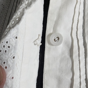 May include: Close-up of a white garment with a button closure. The fabric has vertical pleats and a decorative eyelet lace trim. A black stripe runs along the button line. The button is round and white.