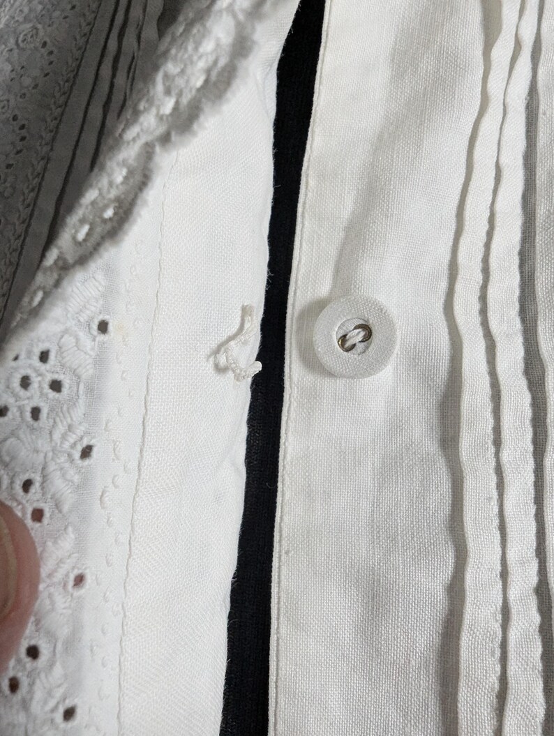 May include: Close-up of a white garment with a button closure. The fabric has vertical pleats and a decorative eyelet lace trim. A black stripe runs along the button line. The button is round and white.
