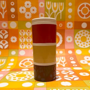 Könnte beinhalten: Ein stapelbarer Behälter im Retro-Stil mit weißem Deckel und drei Abschnitten in Rot, Gelb und Braun. Der Behälter steht vor einem farbenfrohen Hintergrund mit orangefarbenen, gelben und weißen Blumen- und geometrischen Mustern.