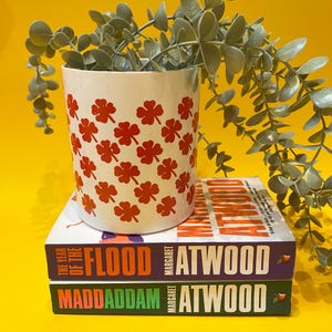 May include: A white ceramic planter with a red clover pattern, holding a green plant, sits atop two books by Margaret Atwood. The books are titled "The Year of the Flood" and "MaddAddam". The background is yellow.