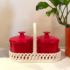 May include: A set of two bright red ceramic containers with matching lids, presented on a white woven tray with a handle. The containers are cylindrical and have a glossy finish. A houseplant in an orange pot is in the background.